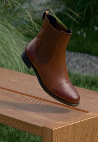Brown leather Chelsea boot with perforated cap toe and elastic side panels, featuring a low heel and smooth finish, floating above a wooden surface.