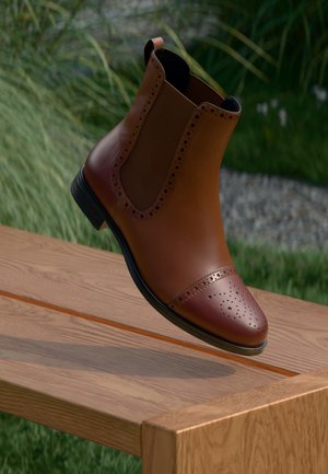 Brown leather Chelsea boot with perforated cap toe and elastic side panels, featuring a low heel and smooth finish, floating above a wooden surface.