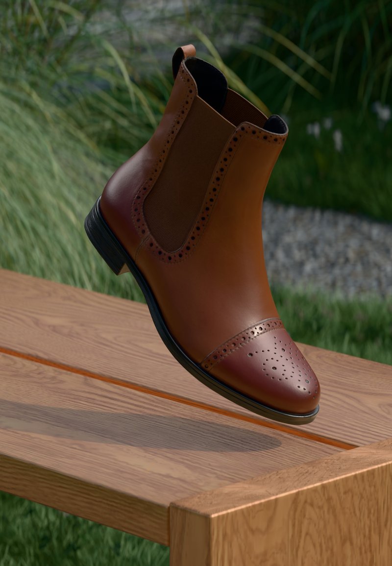 Brown leather Chelsea boot with perforated cap toe and elastic side panels, featuring a low heel and smooth finish, floating above a wooden surface.