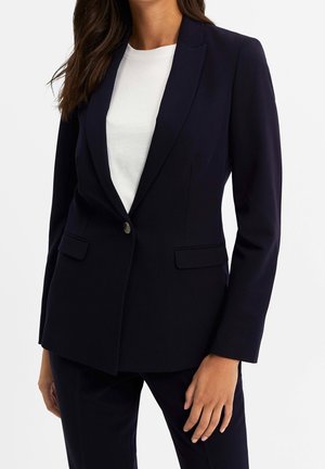 Femme portant un blazer bleu marine foncé avec un seul bouton sur un haut blanc, assorti à un pantalon bleu marine foncé.