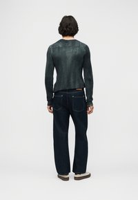 Άτομο με κοντά σγουρά μαλλιά, που φοράει σκούρο πράσινο πλεκτό πουλόβερ με ριγέ υφή, σκούρο μπλε τζιν παντελόνι και μπεζ αθλητικά παπούτσια, στέκεται με την πλάτη προς το μέρος μας σε λευκό φόντο.