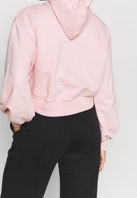 Sudadera corta de color rosa claro con capucha, dobladillo acanalado y mangas globo, hecha de tela suave, mostrada desde atrás sobre un fondo neutro.