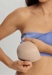 Beige Silikonen-BH-Cup mit glatter Textur, in einer Hand gehalten, neben einem blauen, gerippten BH-Oberteil. Subtile Krümmung für eine gute Passform.