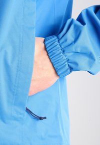 Blå tygjacka med en texturerad yta, med elastiska ärmslut och en sidoficka, detaljerad med justerbar dragsko.