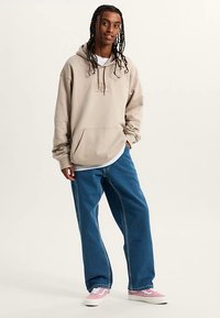 Beige luvtröja med huva och framficka, använd över en vit skjorta. Blå pösiga jeans och rosa-vit-sneakers kompletterar outfiten.