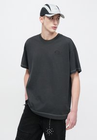 Sötétszürke, bő fazonú pamut póló kerek nyakkal és kontrasztos varrással, fekete, kopott hatású farmerral, szegecses díszítésekkel párosítva.