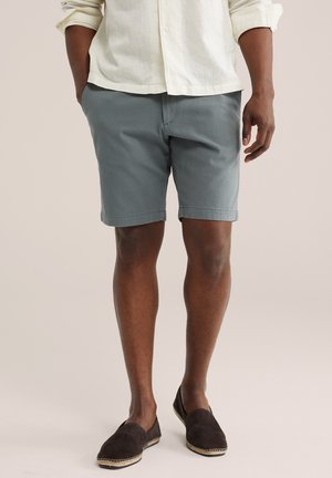 Mann trägt ein hellbeiges Langarmshirt, graue maßgeschneiderte Shorts und dunkelbraune Slipper, steht vor einfachem Hintergrund.