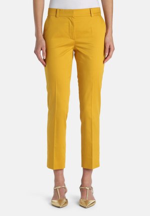 Femme portant un pantalon sur mesure moutarde longueur cheville et des escarpins dorés à brides en T à bouts pointus sur fond blanc.