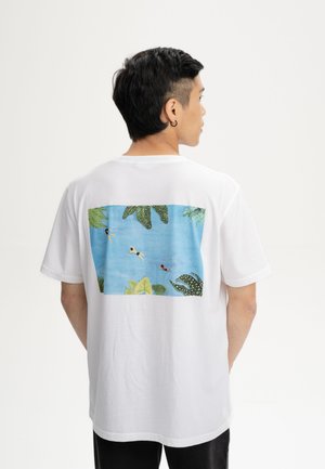 Mann trägt weißes T-Shirt mit Rückendruck von Schwimmern in blauem Wasser, umgeben von grünen Blättern.