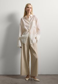 Blusa de satén blanca con mangas largas y un bolsillo en el pecho, combinada con pantalones de lino beige de pierna ancha y tacones puntiagudos color crema.