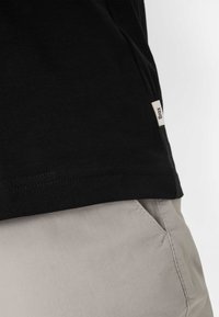 Zwart T-shirt met ribbeltextuur, voorzien van een klein logo-label aan de zijkant. Gecombineerd met lichtgrijze broeken. De nadruk ligt op de stof- en kleurdetails.