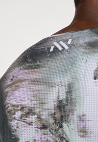 Kurzärmliges gemustertes Hemd mit einem Gittertextur, abstrakten Farben in Grau und Lila, und einem weißen Logo auf dem Rücken.