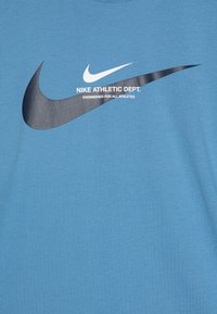 Zila kokvilnas T-kreklu ar balto un melno Nike swoosh logotipu, ar tekstu "Nike Athletic Dept." un "Izstrādāts visiem sportistiem."