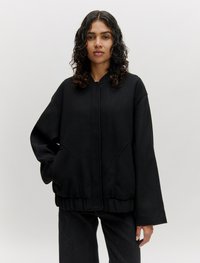 Zwarte oversized hoodie van gestructureerde stof, met een voorste rits, grote zakken en brede mouwen. Versterkt met een geribbelde zoom.