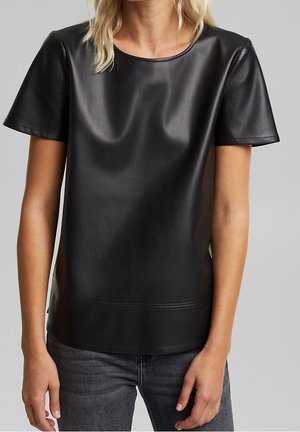 Kobieta ubrana w czarną, krótkim rękawem, sztuczną skórzaną bluzkę w połączeniu z ciemnoszarymi jeansami na tle jednolitego szarego tła.