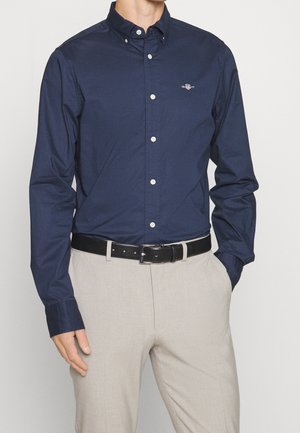 Man wearing navy blue button-up shirt tucked into light beige pants with black leather belt, hand in pocket, against plain background.