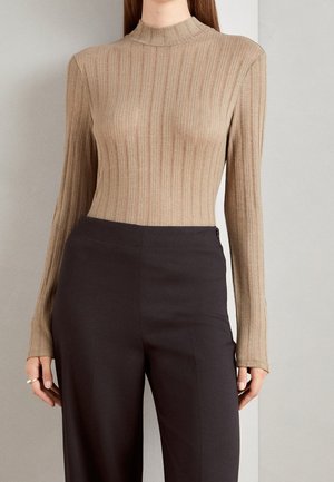 Vrouw draagt een aansluitende beige geribde coltrui en loszittende zwarte broek met hoge taille, staand tegen een effen lichtgrijze achtergrond.