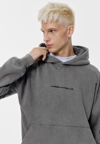 Szara bluza z kapturem z przednią kieszenią typu kangur, z haftowanym napisem "Założono w Nowym Jorku, 2025" oraz ściągaczami przy mankietach i dolnym brzegu. Miękka tekstura.