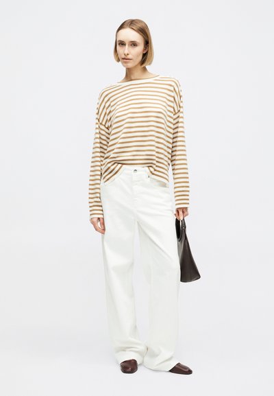 Femme portant un haut à manches longues beige et blanc rayé, un pantalon blanc à jambes larges, des chaussures marron, et tenant un sac à main marron foncé.