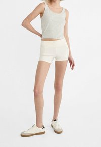 Osoba w jasnoszarym podkoszulku, białych dopasowanych szortach i białych trampkach z gumowymi podeszwami, stojąca na tle o białym kolorze.