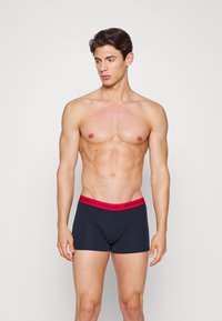 Homme torse nu portant un boxer noir avec une ceinture rouge portant l'inscription "Tommy Hilfiger", se tenant devant un fond clair.