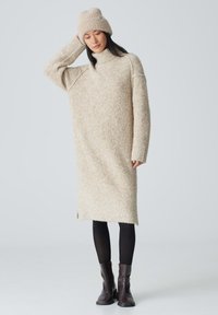 Beige, übergroßes Strickkleid mit Stehkragen, langen Ärmeln und seitlichen Schlitzen. Kombiniert mit schwarzen Leggings und braunen Stiefeletten.