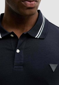 Granatowa koszulka polo z bawełny, z dwoma guzikami, kołnierzykiem w białe i zielone paski oraz trójkątną naszywką z logo na piersi.