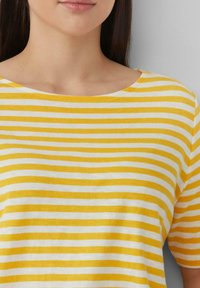Gelb-weiß gestreiftes Shirt mit weitem Halsausschnitt, kurzen Ärmeln und einer weichen Textur, das auf einer Person von den Schultern aufwärts zu sehen ist.