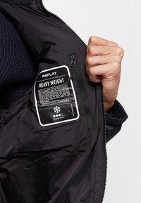 Zwarte gewatteerde jas met een opvallend etiket waarop "REPLAY HEAVY WEIGHT" staat. Beschikt over een ritszak en een gladde, glanzende textuur.