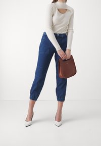 Bílý žebrovaný top s dlouhým rukávem a výřezem na krku, v kombinaci s užšími modrými džíny a bílými lodičkami na špičce, držící hnědou kabelku.