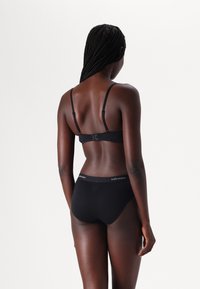 Completo di lingerie nera con una texture morbida e liscia. Include un reggiseno semplice con spalline regolabili e un perizoma coordinato con cintura con logo.