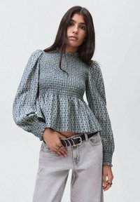 Blusa blu con motivo, dotata di corpetto arricciato e maniche lunghe a sbuffo, abbinata a jeans grigi chiari a vita alta e una cintura nera.