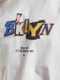 Balta marškinėliai su spalvinga, mišria tipografija, rašančia "BKLYN", o žemiau juodu tekstu – "SPREAD LOVE IT'S THE BROOKLYN WAY 1994."