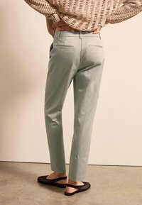 Hellgrüne maßgeschneiderte Hose mit einer glatten Textur, ausgestattet mit zwei Gesäßtaschen, kombiniert mit schwarzen, offenen Mesh-Sandalen auf einem Betonboden.