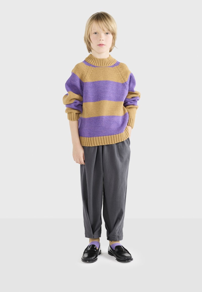 Maglione a maglia con strisce viola e senape, colletto e polsini a costine; abbinato a pantaloni grigi larghi e mocassini neri. Calze viola visibili.