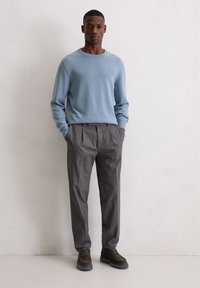 Jasnoniebieski dziergany sweter z długimi rękawami w zestawieniu z szarymi plisowanymi spodniami. Ciemne buty dopełniają strój, na tle neutralnego tła.