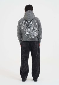 Persona che indossa una giacca grigia con cappuccio decorata con motivi floreali e tigri, abbinata a pantaloni neri con stampe di serpenti scuri e teschi, in posizione di spalle.