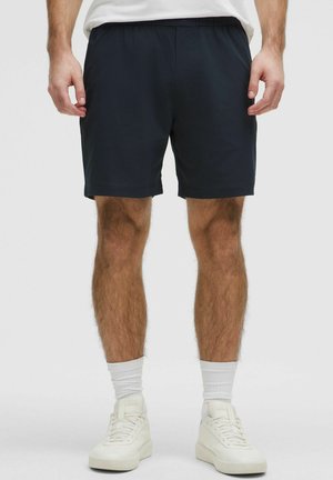 Hombre vistiendo pantalones cortos deportivos negros, calcetines blancos hasta la mitad de la pierna y zapatillas blancas, posando contra un fondo neutro.