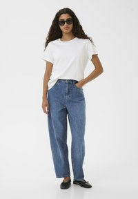 T-shirt bianca a maniche corte, jeans in denim blu a vita alta e scarpe nere flat. Modella con occhiali da sole neri. Sfondo semplice.