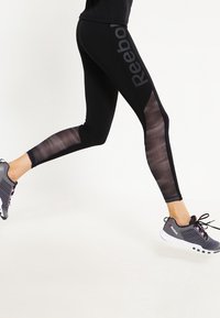 Czarne legginsy sportowe z siateczkowymi wstawkami po bokach, z logotypem Reebok w wyraźnym szarym kolorze. Połączone z szarymi butami sportowymi.