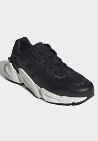 Zapatilla deportiva negra con parte superior de malla y sintético, acentos negros texturizados, suela intermedia blanca gruesa y sistema de cordones tradicional.
