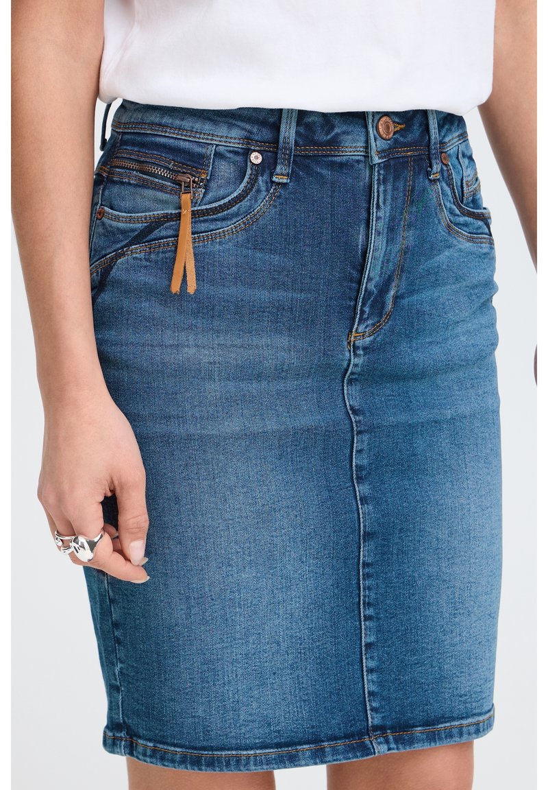 Personne portant une jupe en jean bleu et un haut blanc, avec un détail de tirette marron et des bagues argentées à la main gauche, debout devant un fond uni.