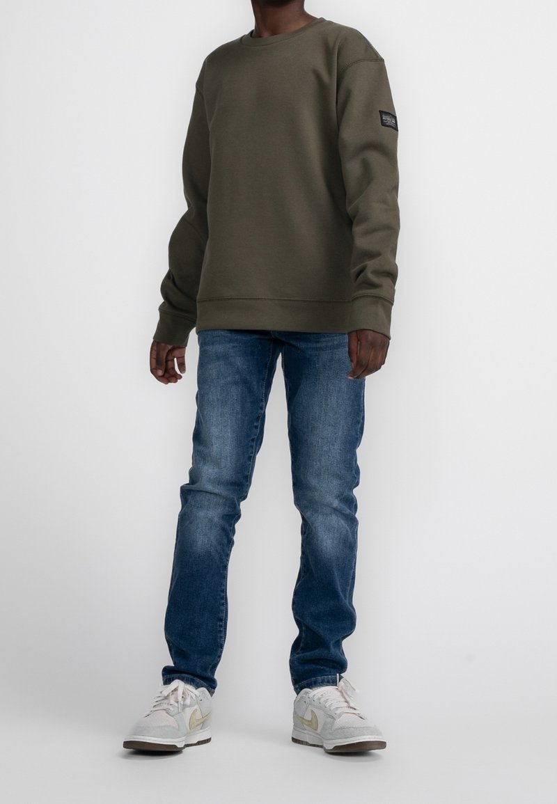 Personne portant un sweat-shirt vert olive, un jean bleu et des baskets blanches, se tenant devant un fond clair uni.