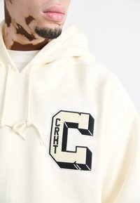 Sudadera crema con una prominente letra "C" bordada en negro y beige, y el texto "CRHT". Cuenta con capucha con cordón y una textura de algodón suave.