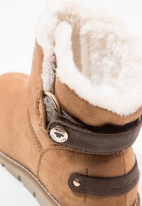 Bottine en daim marron avec une doublure en fausse fourrure, dotée d'une sangle en cuir avec un détail bouton en métal et une semelle plate en caoutchouc.