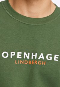 Felpa di cotone verde con la scritta bianca "OPENHAGE" e arancione "LINDBERGH" sulla parte anteriore. Girocollo con dettagli a coste.