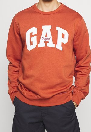 Mann trägt einen orangefarbenen GAP-Sweatshirt mit weißen Buchstaben und rotem "Original"-Text, kombiniert mit dunkler Hose, Hände in den Taschen, vor einfachem Hintergrund.