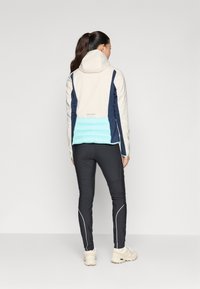 Lys beige jakke med marineblå og turkise seksjoner, med hette og quiltet nedre del av ryggen; parret med svarte tettsittende bukser og beige sneakers.