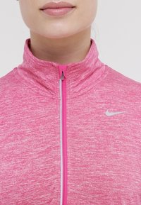 Nahaufnahme einer Person, die eine pinke, melierte Nike-Jacke mit Reißverschluss und hohem Kragen trägt, mit reflektierendem Logo auf der Brust.