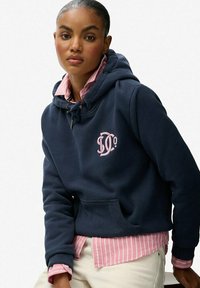 Mujer con el pelo recogido lleva una sudadera azul marino sobre una camiseta de rayas rosas y pantalones claros, sentada y mirando hacia adelante con una expresión neutral.